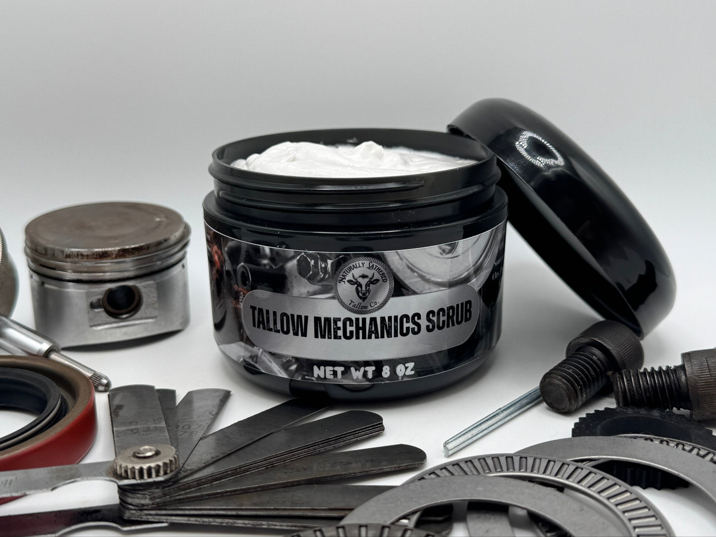 Jar of 'Tallow Mechanics Scrub' surrounded by mechanical parts on a light gray background